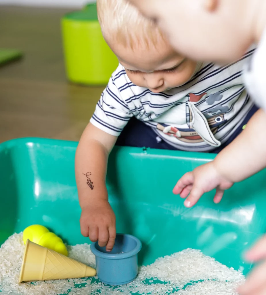 Deux enfant dont un petit garçon blond jouent avec un bac de motricité dans une micro-crèche.